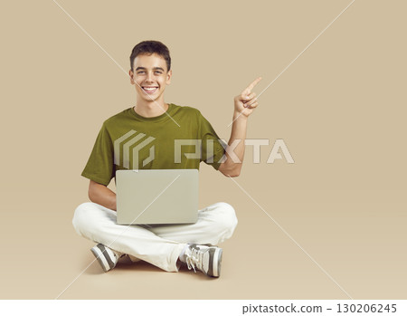 Happy teenage student boy with laptop computer pointing finger away on beige background Happy teenage student boy with laptop computer pointing finger away on beige background 130206245