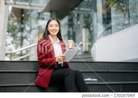 Confident Asian woman with a smile standing holding notepad and tablet at out side office. Confident Asian woman with a smile standing holding notepad and tablet at out side office. 130207069