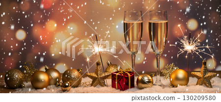 Christmas celebration with champagne glasses, sparklers, golden baubles, and red gift on snow with warm festive background 130209580