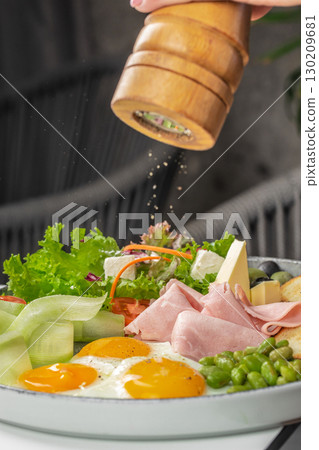 Fresh breakfast plate with fried eggs, ham, vegetables, and cheese being seasoned with pepper 130209681