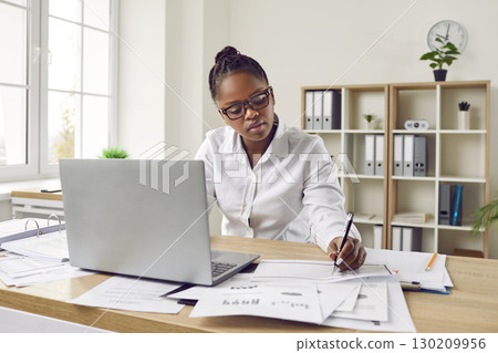 Serious, focused african-american girl with glasses is sitting at her desk in front of a laptop monitor in the office. 130209956