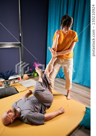 Masseuse making thai yoga massage. Woman therapist doing traditional massage treatment using elbow for male patient. Bald client lying on mat, meditating, enjoying hand massage in calm atmosphere. 130210924