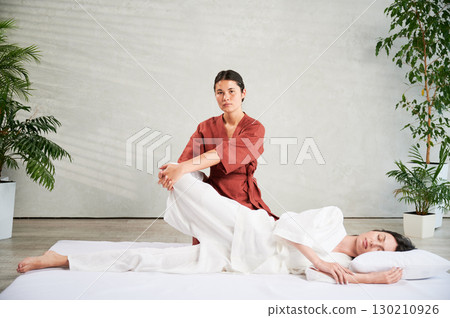 Masseuse making thai yoga massage. Woman therapist doing traditional massage treatment, stretching female patient's body. Client lying on mat, enjoying hand massage in calm atmosphere. 130210926