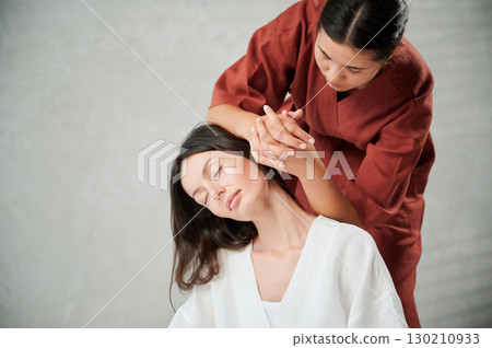 Masseuse making thai yoga massage. Woman therapist doing traditional massage treatment for female patient indoors. Client sitting, meditating, enjoying hand massage in calm atmosphere. 130210933