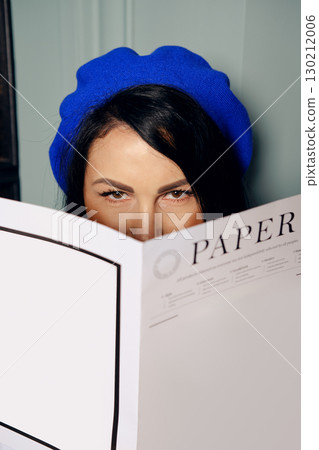 Woman in blue beret reading a large paper while peeking over it indoors 130212006