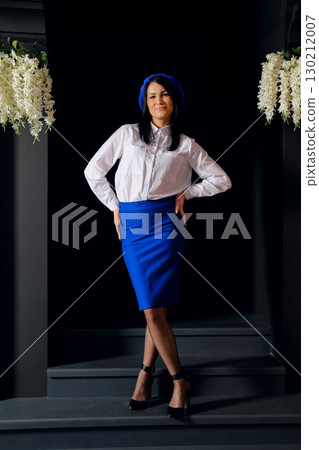 Elegant woman poses confidently in a stylish blue outfit with a chic hat against a dark backdrop 130212007