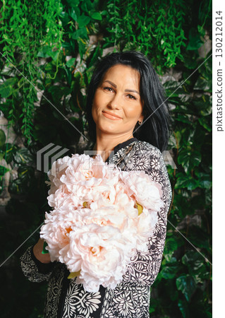Woman holding large bouquet of pink flowers in a lush green indoor setting during daylight 130212014
