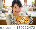 A woman wearing an apron holds a freshly baked apple pie and has a warm smile in a homely setting 130212472