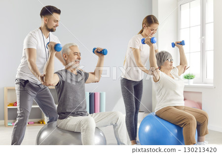 Health care workers helping mature people to do sport exercises with dumbbells sitting on fit ball in gym. Health care workers helping mature people to do sport exercises with dumbbells sitting on fit ball in gym. 130213420
