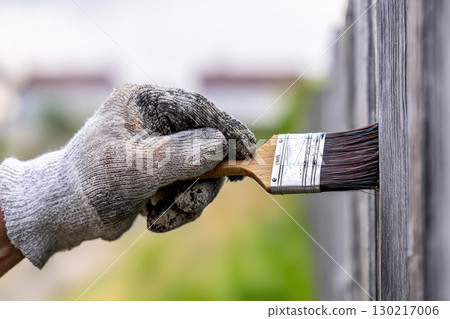 Hand applying deep brown paint to a textured wooden fence plank in daylight for a fresh look and renewed protection 130217006