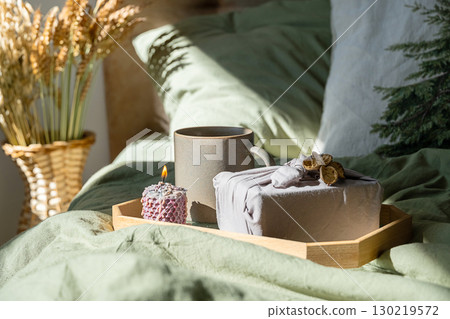 Cozy bed scene with mug, lit candle and furoshiki gift on wooden tray. Cozy bed scene with mug, lit candle and furoshiki gift on wooden tray. 130219572
