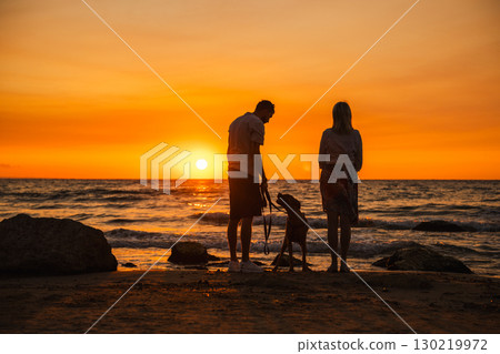 Couple with a German Boxer dog standing on the sandy shore, gazing at the glowing sunset over the sea horizon during summer evening walk. 130219972