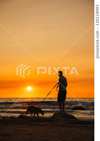 Man standing on a rocky shore holding a German Boxer dog on a leash as the dog explores near the water during a sunset by the sea. 130219993