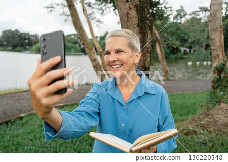 Happy senior woman video calling while holding a book in a picturesque park by the lake Happy senior woman video calling while holding a book in a picturesque park by the lake 130220548