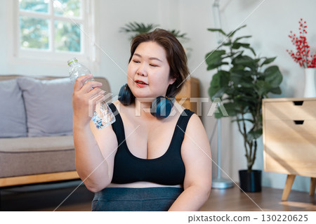 Plus-size woman holding a water bottle, reflecting on hydration and healthy lifestyle choices at home 130220625