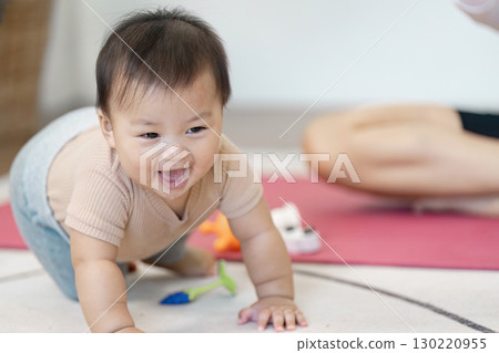 Joyful crawling baby exploring toys during playtime, with a multitasking mother nearby at home 130220955