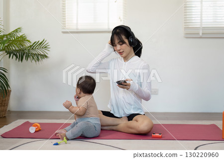Multitasking mother enjoying music on her phone while keeping an eye on her baby during playtime at home 130220960
