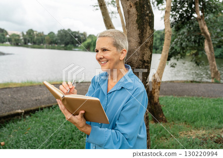 Joyful senior woman writing in a notebook while enjoying the peaceful lakeside view in a park Joyful senior woman writing in a notebook while enjoying the peaceful lakeside view in a park 130220994