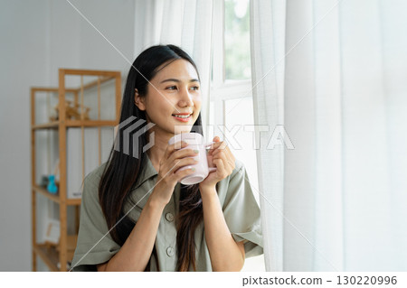 Smiling woman enjoying a cup of coffee by the window in a cozy and bright home setting Smiling woman enjoying a cup of coffee by the window in a cozy and bright home setting 130220996