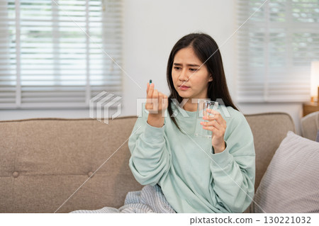 Woman in a mint green sweater thoughtfully examining a pill while holding a glass of water at home, preparing for relief from discomfort Woman in a mint green sweater thoughtfully examining a pill while holding a glass of water at home, preparing for relief from discomfort 130221032
