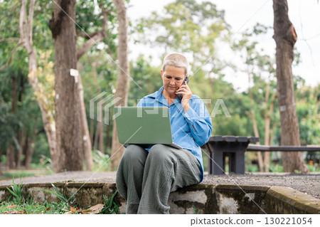 Senior woman engaged in a phone call while working on her laptop in a park 130221054