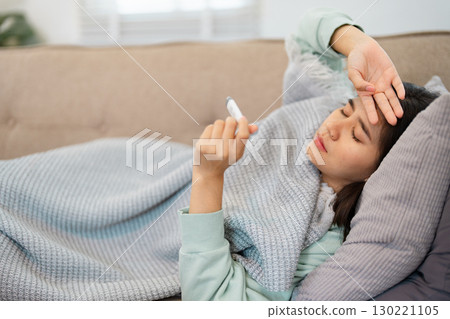 Woman in a mint green sweater lying on a sofa at home, checking her temperature with a thermometer, exhibiting signs of sickness 130221105