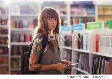 Portrait of young Caucasian woman with tattoo wearing backpack shopping in bookstore. Student searching textbook. Concept of education 130225698