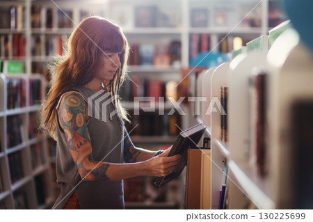 Side view of young pretty Caucasian woman with tattoo choosing books in bookstore. Copy space. Concept of education and shopping in book shop 130225699