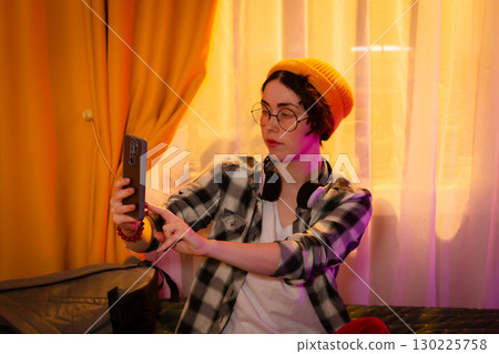 Girl in casual clothes with beanie and headphones relaxing on bed and taking selfie. Sunset glow and cozy home background. Mid shot 130225758