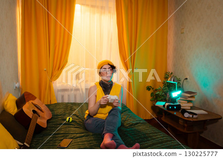 Teen girl enjoying peaceful solo gaming moment in her room. Sitting comfortably on bed, surrounded by warm light. Concept of digital comfort and fun. Wide shot Teen girl enjoying peaceful solo gaming moment in her room. Sitting comfortably on bed, surrounded by warm light. Concept of digital comfort and fun. Wide shot 130225777