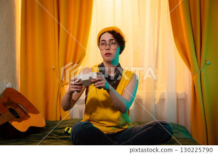 Teenage girl sitting with crossed legs on bed playing games. Tomboy style clothes and colorful room. Concept of indoor leisure and modern culture. Mid shot 130225790