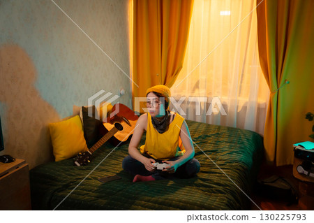 Young gamer girl with glasses sits cross legged on bed with game controller. Concept of youthful concentration and home comfort. Wide shot, top view Young gamer girl with glasses sits cross legged on bed with game controller. Concept of youthful concentration and home comfort. Wide shot, top view 130225793
