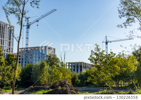 Cranes tower over new buildings surrounded by lush trees and underbrush on a sunny afternoon in a developing urban area Cranes tower over new buildings surrounded by lush trees and underbrush on a sunny afternoon in a developing urban area 130226358