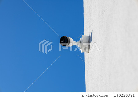 Security camera positioned on a white surface captures the bright blue sky, ensuring surveillance in an open area under sunlight 130226381