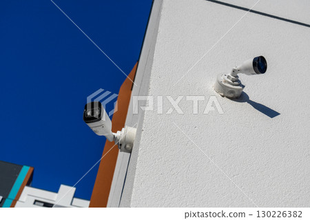 Two security cameras are installed on the corner of a building, watching over the area beneath a bright blue sky during the day 130226382