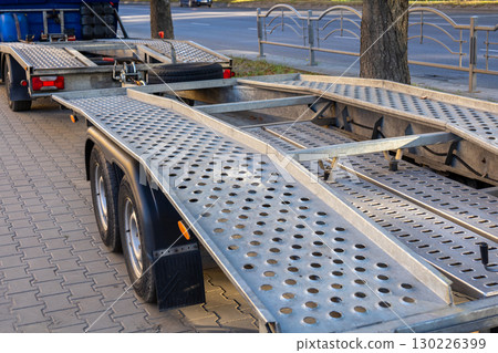 A heavy-duty car hauler sits parked on a city street, displaying its metal ramps and wheels under the afternoon sunlight A heavy-duty car hauler sits parked on a city street, displaying its metal ramps and wheels under the afternoon sunlight 130226399