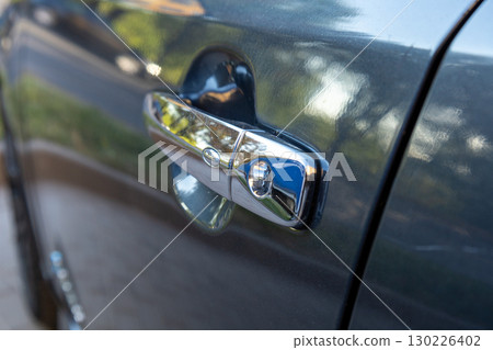 The car door handle reflects sunlight in a suburban neighborhood with blurred greenery in the background, showcasing its chrome finish 130226402