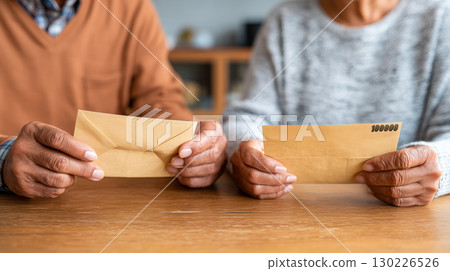 An elderly couple holding an envelope in front of a wooden table indoors | A calm, everyday scene reminiscent of household finances An elderly couple holding an envelope in front of a wooden table indoors | A calm, everyday scene reminiscent of household finances 130226526