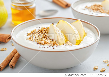 Healthy breakfast bowl with poached pears, creamy yogurt, and crunchy granola set on a neutral table with honey and cinnamon sticks 130227537