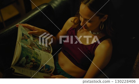 Young woman relaxing on couch reading magazine at night Young woman relaxing on couch reading magazine at night 130228406