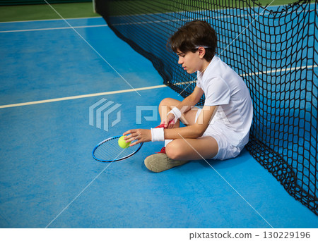 Boy Resting With Tennis Ball and Racket by the Court Net Boy Resting With Tennis Ball and Racket by the Court Net 130229196
