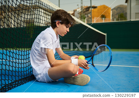 Young Tennis Player Sitting on Court Next to Net Holding Racket Young Tennis Player Sitting on Court Next to Net Holding Racket 130229197