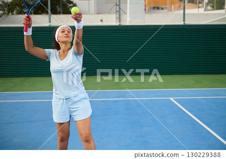 Woman Playing Tennis Focused on Serving Ball Outdoors on Blue Court Woman Playing Tennis Focused on Serving Ball Outdoors on Blue Court 130229388