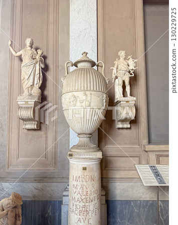VATICAN CITY, ROME, ITALY - AUGUST 02, 2025 Classical marble vase with fluted body and handles sits on an inscribed pedestal. Two smaller statues are on ornate brackets flanking the central urn 130229725