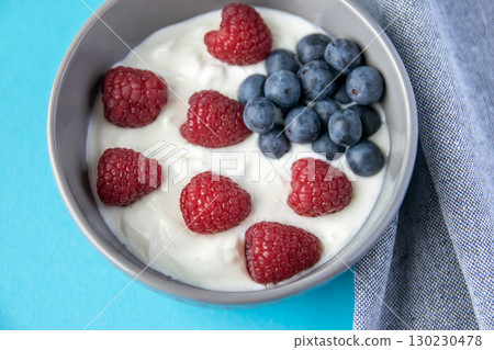 Natural fresh greek yogurt with red raspberry, blue berries in the grey plate and napkin for healthy breakfast. Blue paper background. Natural fresh greek yogurt with red raspberry, blue berries in the grey plate and napkin for healthy breakfast. Blue paper background. 130230478