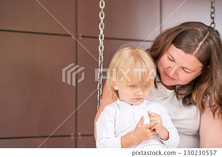Caucasian female adult and young child bonding on swing in warm indoor setting Caucasian female adult and young child bonding on swing in warm indoor setting 130230557