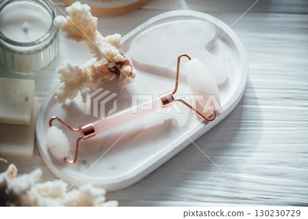 Rose quartz face roller and gua sha stone on a white ceramic tray with coral decor and a lit candle in soft natural light for spa and skincare themes.. Rose quartz face roller and gua sha stone on a white ceramic tray with coral decor and a lit candle in soft natural light for spa and skincare themes.. 130230729