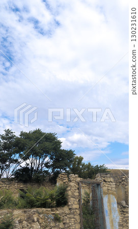 Crumbling stone wall with rusty metal door frame standing beneath cloudy blue sky 130231610