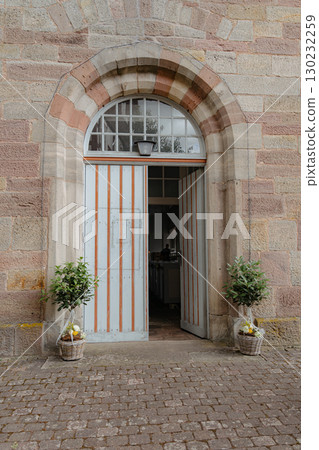 Stone archway with open wooden doors adorned with potted plants in baskets. Captures the charm of historic architecture with inviting entrance and traditional design elements. 130232259