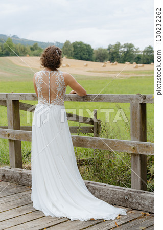 Bride in a flowing white dress stands on a rustic wooden bridge, gazing at a picturesque countryside view with fields and trees in the background, capturing a serene and thoughtful moment Bride in a flowing white dress stands on a rustic wooden bridge, gazing at a picturesque countryside view with fields and trees in the background, capturing a serene and thoughtful moment 130232262